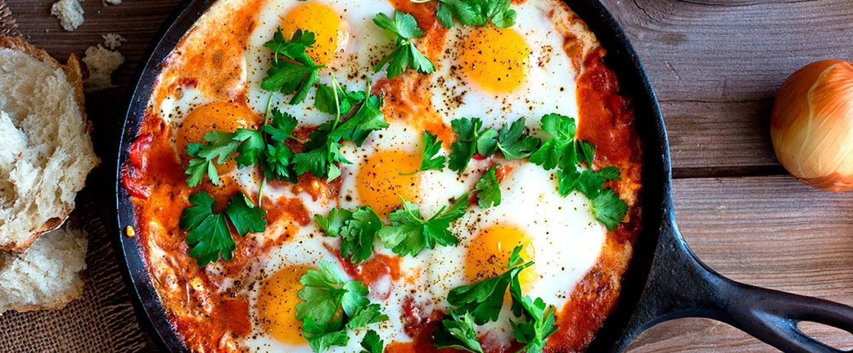 Shakshuka colorée à la sauce tomate et aux œufs, parsemée d'herbes, servie dans une poêle sur un support en bois.